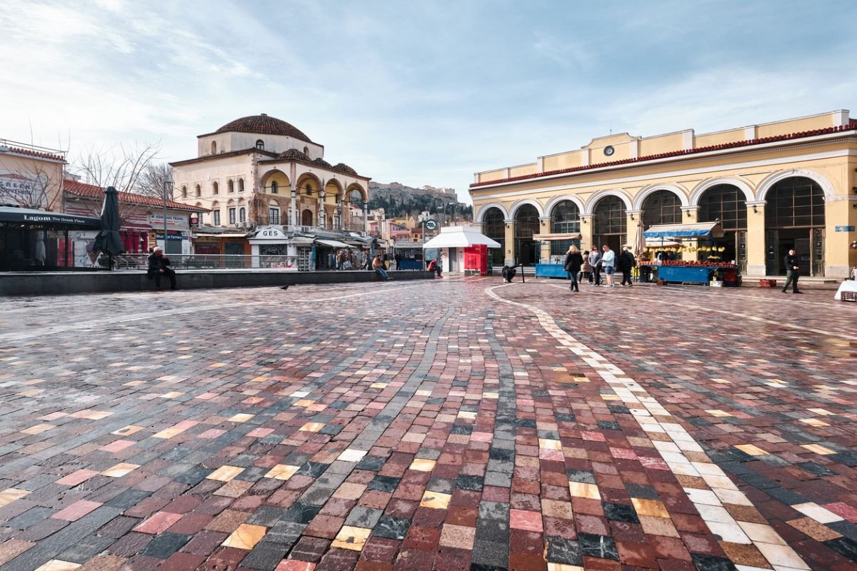 A nearly empty square in Monastiraki, Athens, showcasing colorful cobblestone pavement. The scene includes historical buildings, such as a mosque with a domed roof and arches on the left, and a neoclassical building with arched windows on the right. A few people are walking or sitting in the area, with the Acropolis visible in the background. The sky is overcast, giving the square a calm and serene atmosphere.